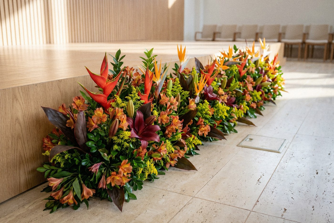 A composição longa correndo rente ao chão no pé do palco: helicônias vermelhas e aves-do-paraíso laranja saindo de uma base densa de alstroemérias, lírios vinho, solidago e folha de cordyline escura, sobre piso de travertino claro, contra a frente de carvalho claro do palco e a parede de ripado de madeira do salão.